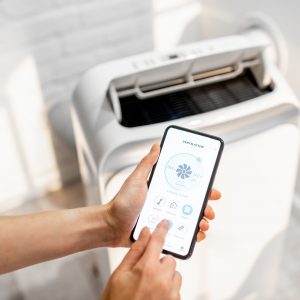 Woman holding smart phone with launched application for air condition control with ventilation unit on the background. Smart home concept